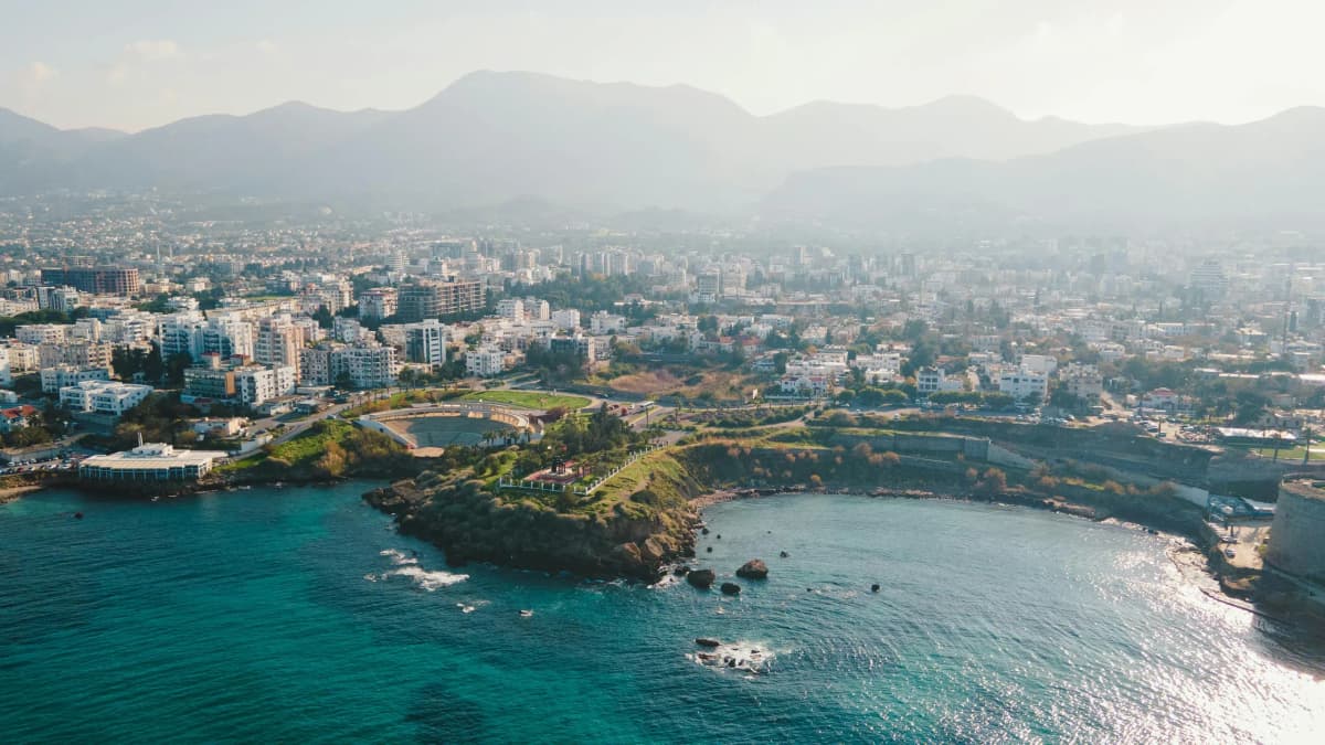 Alt Text: Aerial view of Limassol, Cyprus: urban skyline with white buildings, green peninsula in turquoise bay, distant mountains.