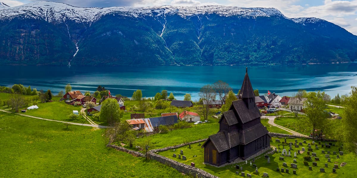 Panoramic view of a Norwegian fjord with turquoise waters, snow-capped mountains, a small village, and the historic Urnes Stave Church in the foreground, representing Norway’s landscape and heritage within a modern digital economy.